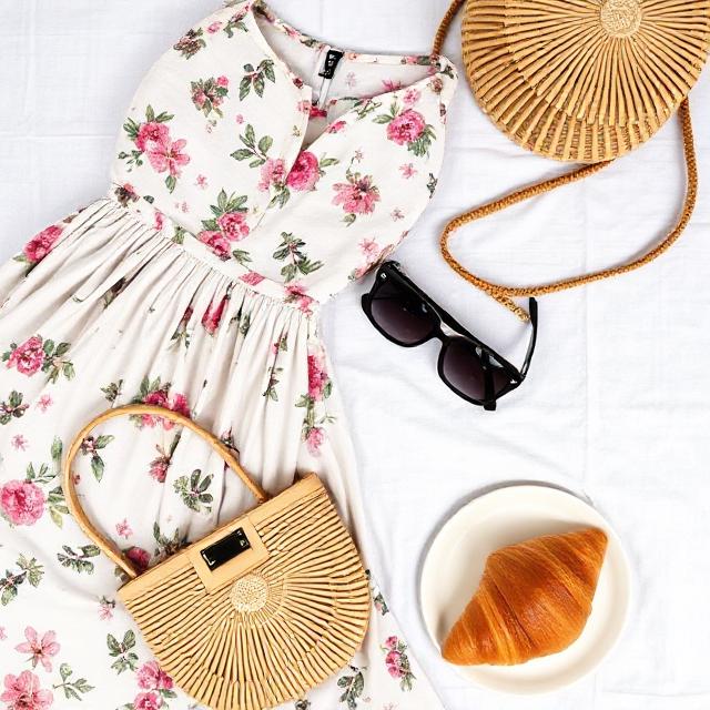 Flat lay of floral dress, woven bag, sunglasses, and croissant on white linen